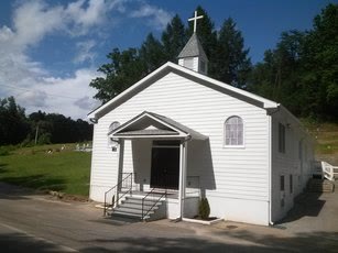 The white clapboard church with a small steeple and cross, a covered front entrance, and wooded hills behind it.