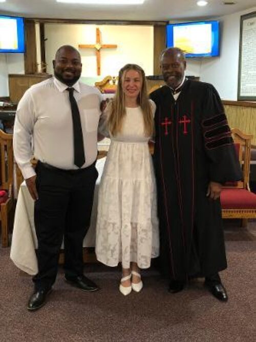Deacon and Deaconess Trammell stand with Pastor Harbin, in his robe, at the front of the sanctuary.
