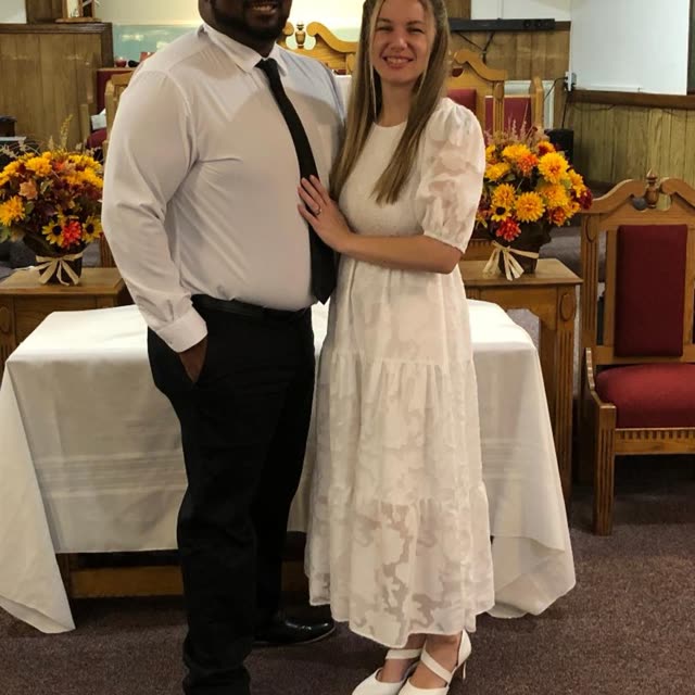A man in a white shirt and tie and a young woman in a white dress stand in the sanctuary.
