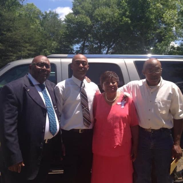 Men in suits and a woman in pink stand together outside beside a car.