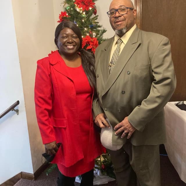 A woman in a red dress and a man in a suit stand by a Christmas wreath.
