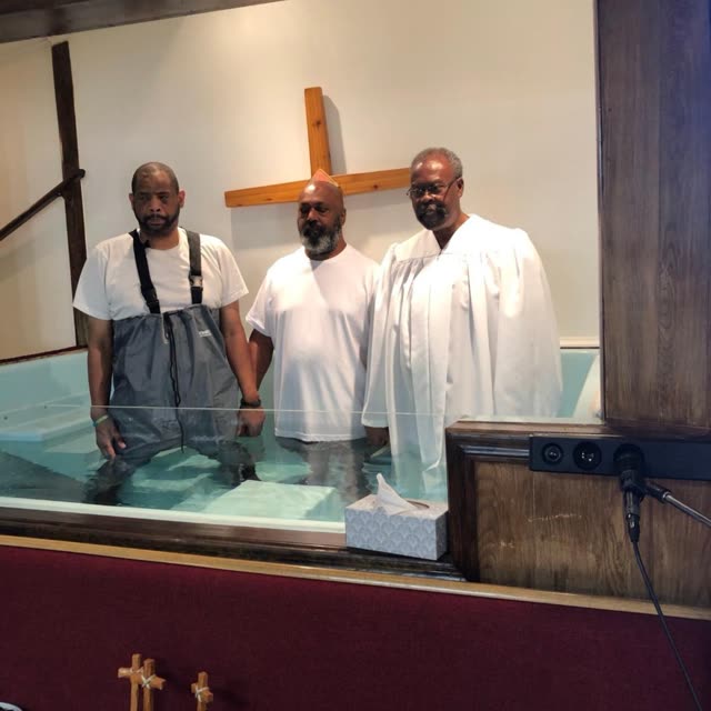 Three men dressed in white stand at the front of the sanctuary under the cross.
