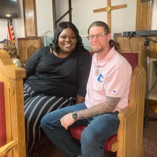 A woman and a man in a pink shirt sit together in a pew.