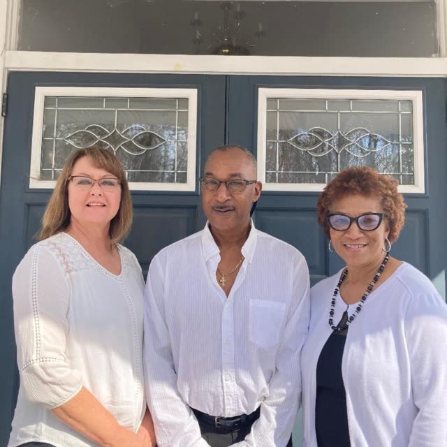 Two women and a man dressed in white stand in front of the church doors.