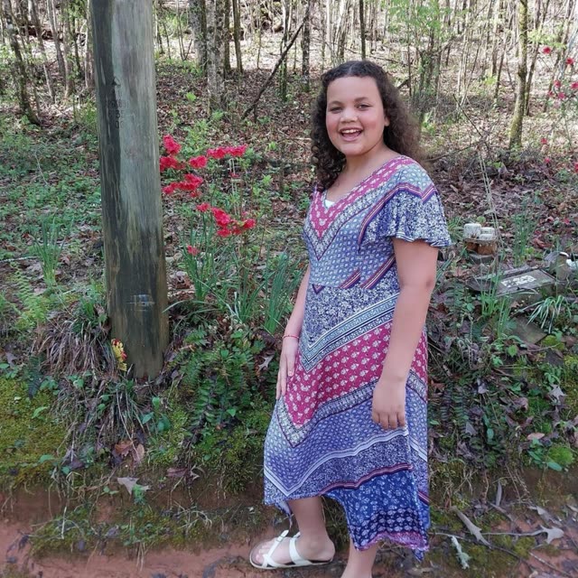 A woman in a patterned dress stands outdoors among trees.