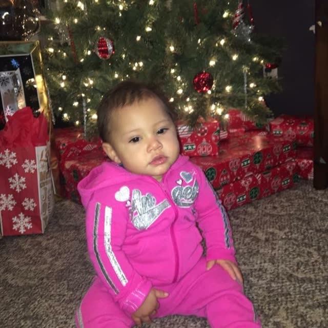 A toddler in a pink outfit stands by a Christmas tree.