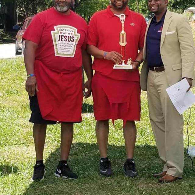 Two men in red shirts and a man in a suit stand on a lawn.