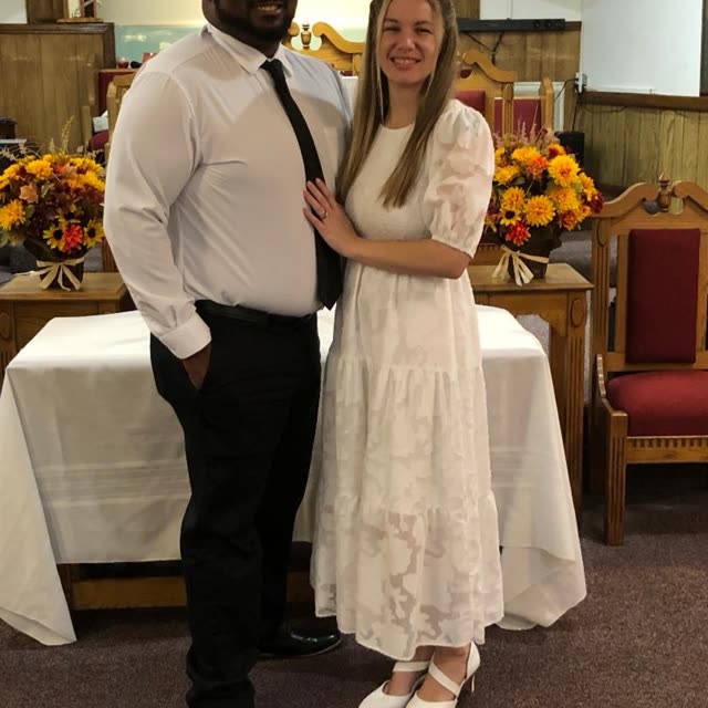 A man in a white shirt and tie and a young woman in a white dress at the front of the sanctuary.