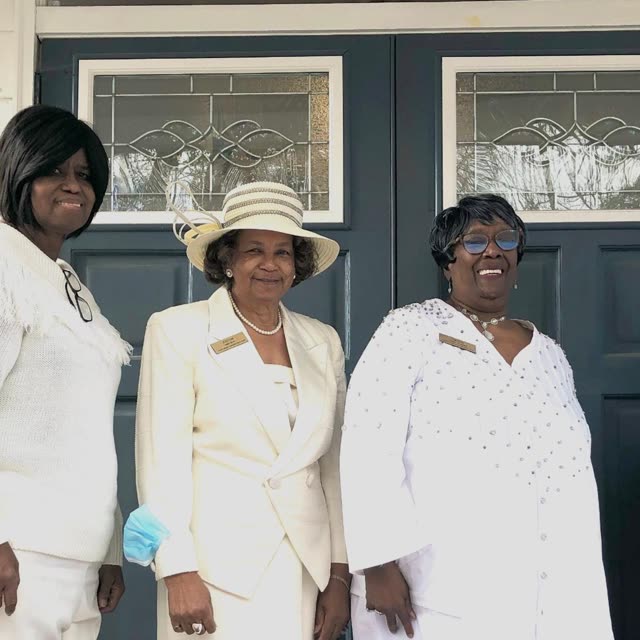 Three women dressed in white, one in a white hat, stand by the church doors.