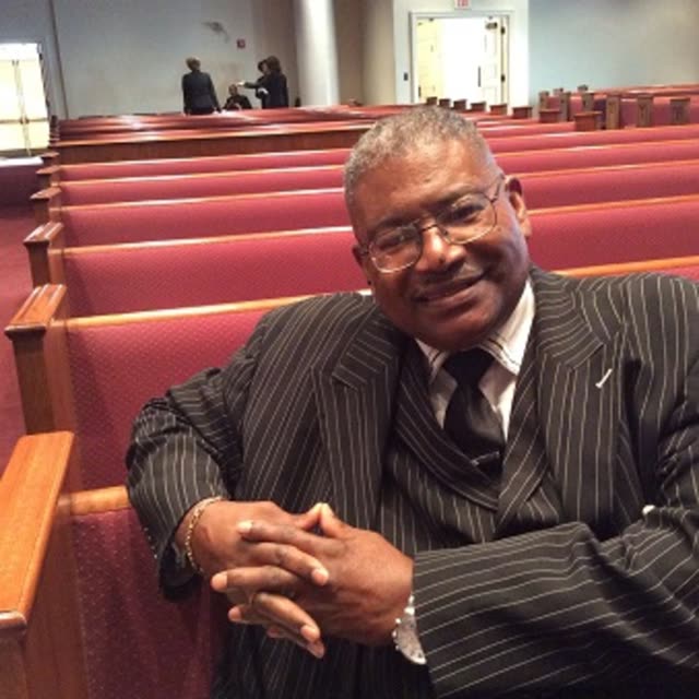 A smiling man in a pinstriped suit sits in a pew.