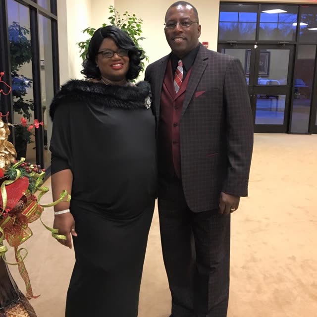 A woman in black and a man in a suit stand together indoors.