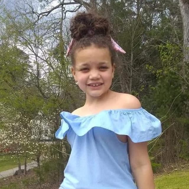 A girl in a blue off-the-shoulder dress stands outdoors.