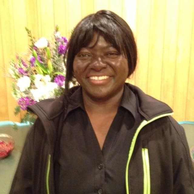 A smiling woman in a dark jacket stands by flowers.