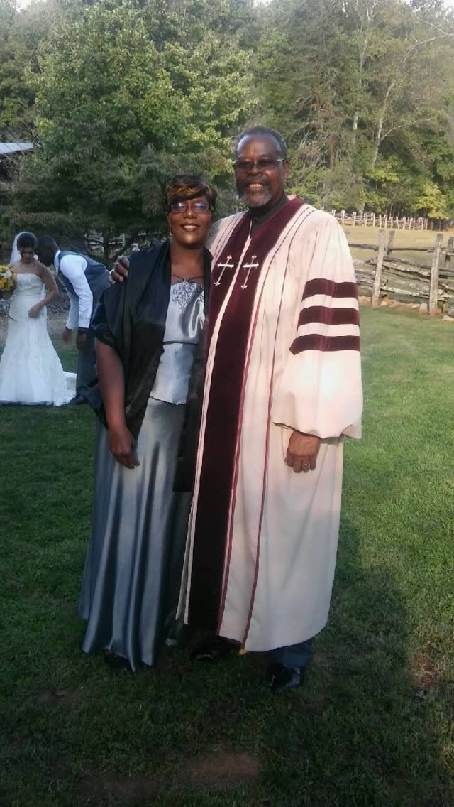A minister in a black robe stands on the lawn with a woman in a long dress.