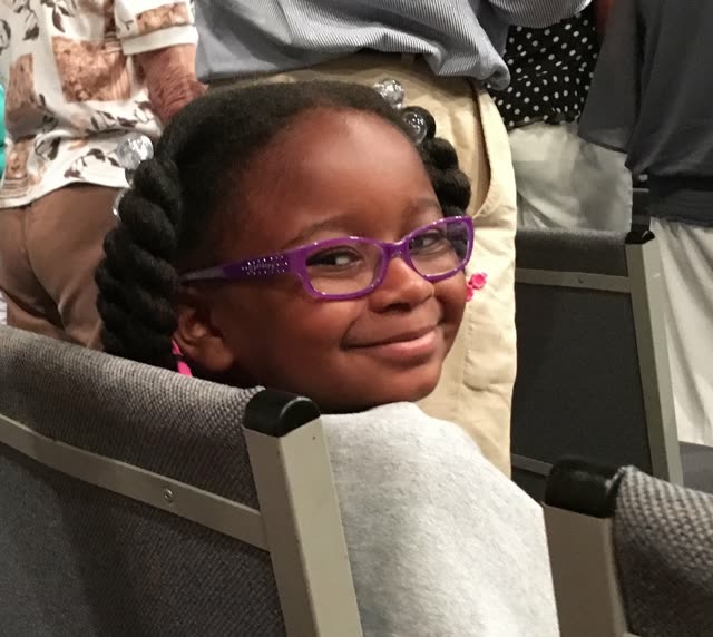 A smiling girl with glasses sits in a church pew.