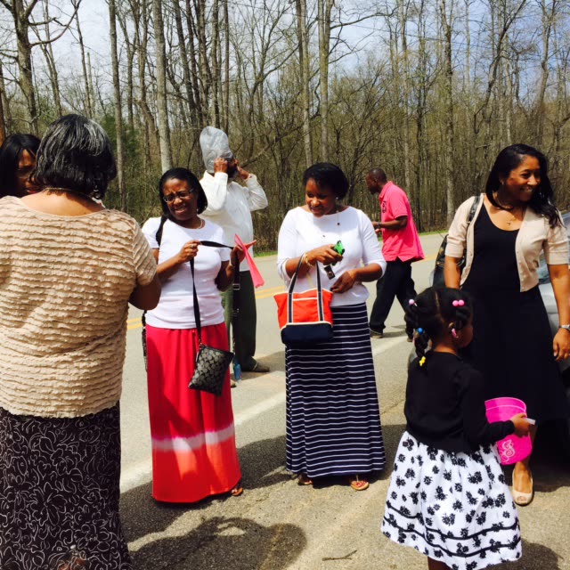 Women and girls in long, colorful skirts gather outdoors among bare trees.