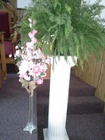 A white pedestal holding a fern and pink flowers.